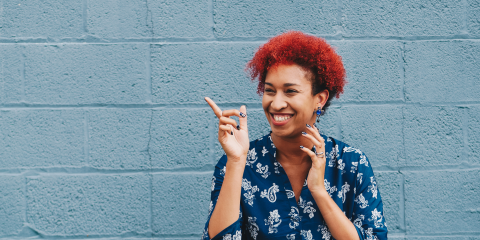 Mulher sorridente com cabelo vermelho e vestido azul em frente a uma parede azul